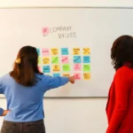 Dua karyawan berdiskusi tentang nilai perusahaan di depan papan tulis penuh catatan berwarna